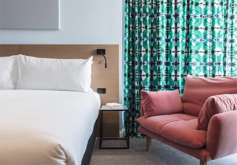 Modern bedroom with a bed, a bedside table, a pink armchair, and a patterned curtain.