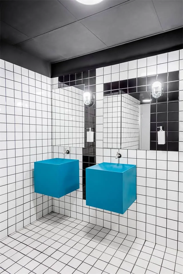 A modern public bathroom features white and black grid-patterned tiles with bright blue square floating sinks and silver fauc
