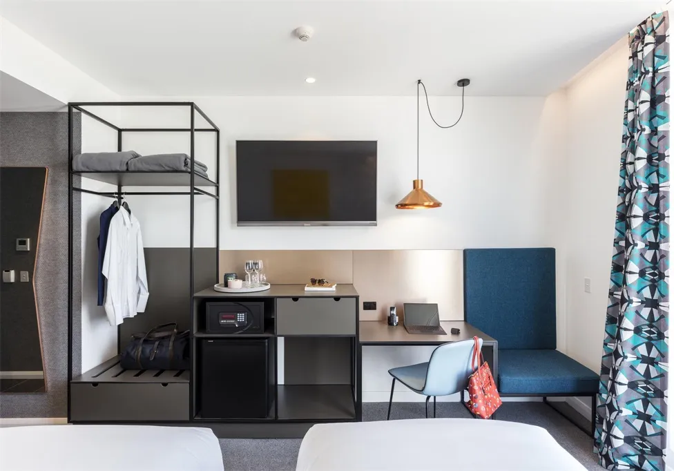 Modern minimalist hotel room with black metal frame cabinet, wooden desk, and fabric seating.