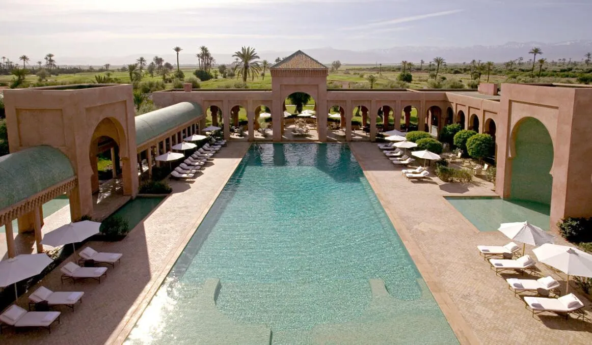 A modern resort with a large swimming pool surrounded by lounge chairs and umbrellas, set against a backdrop of lush greenery