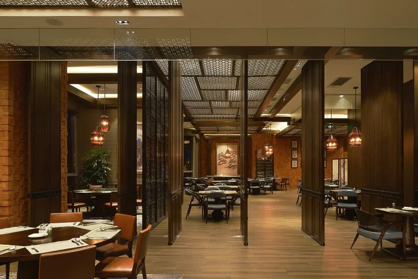 A contemporary restaurant interior with wooden tables, brick walls, and glass pendant lights hanging above round tables.
