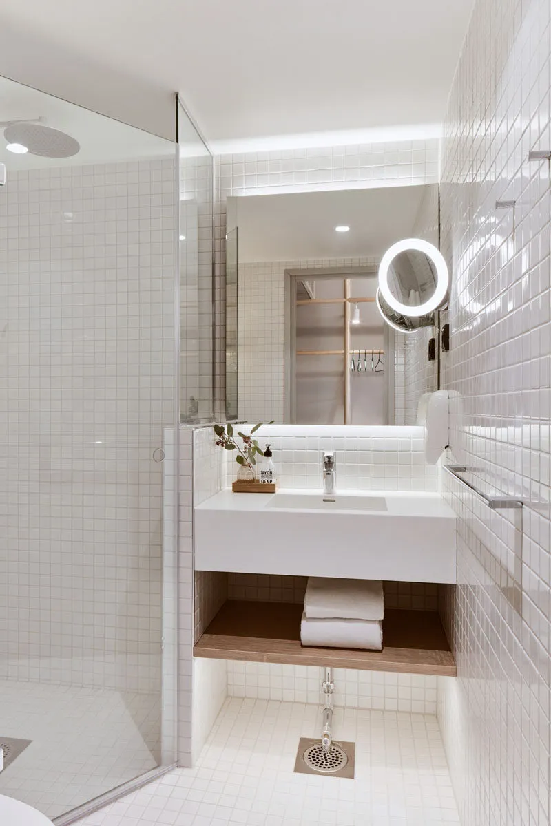 White-tiled bathroom with minimalist design, featuring a sink, shower, and towel storage.