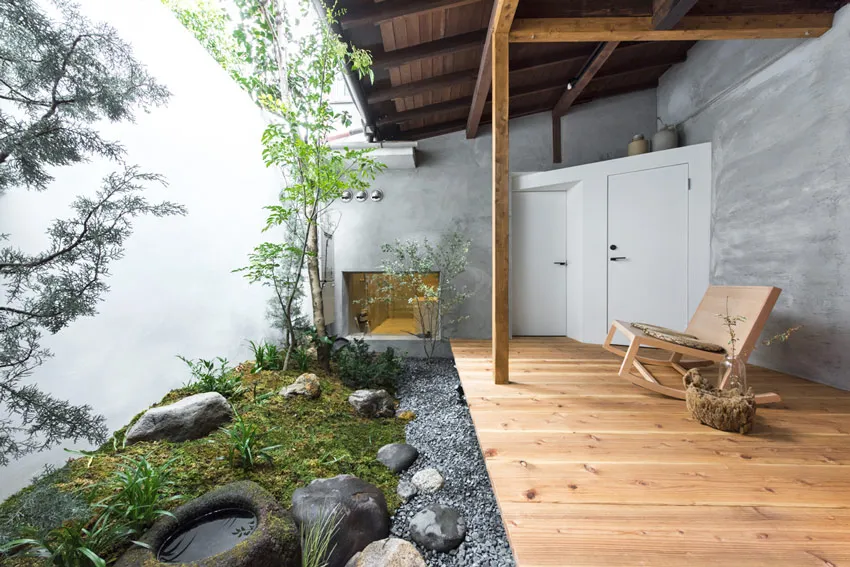 A serene Japanese-style patio with wooden deck, concrete walls, stone and gravel landscaping, and a wooden rocking chair.
