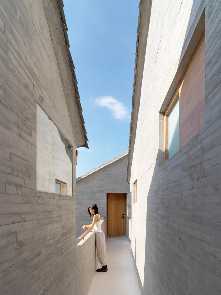 A woman standing in a narrow, concrete-walled corridor with a wooden door and windows.