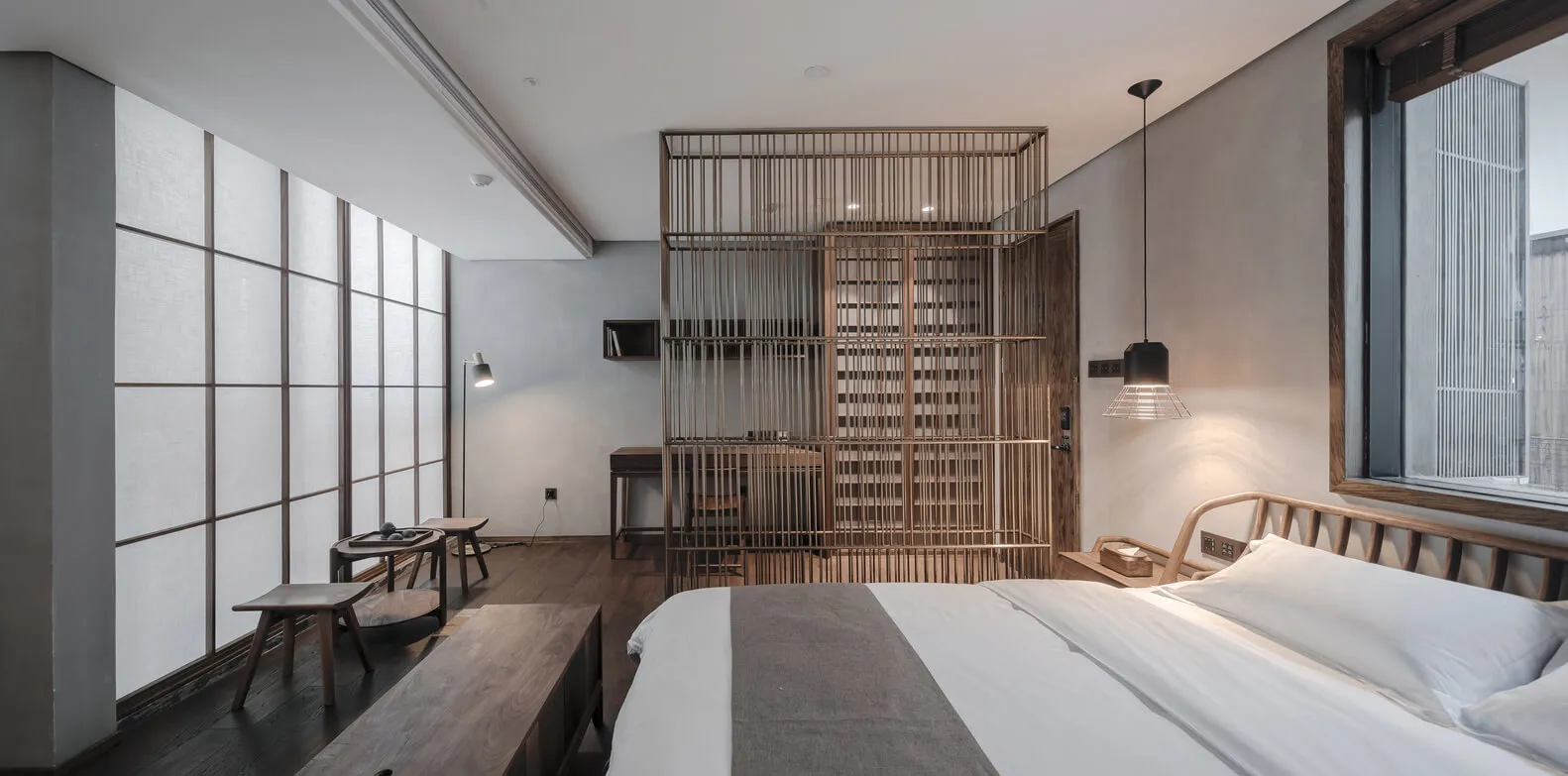 A minimalist bedroom featuring a wooden bed and furniture, with large windows and a hanging lamp.
