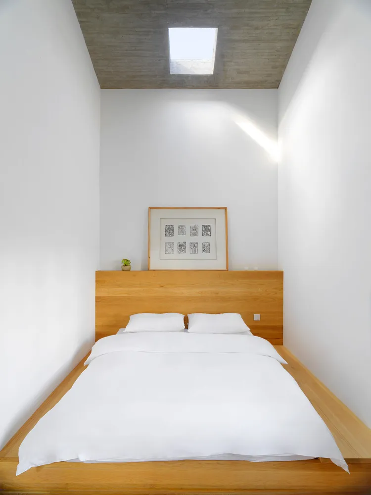 A minimalist bedroom with a wooden platform bed, white linens, and a concrete ceiling with a skylight.