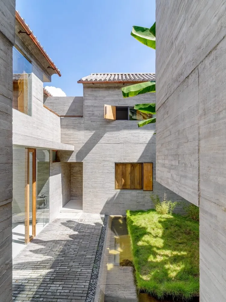 A minimalist house exterior with concrete walls, wooden window shutters, and a small garden with grass and plants, under a cl