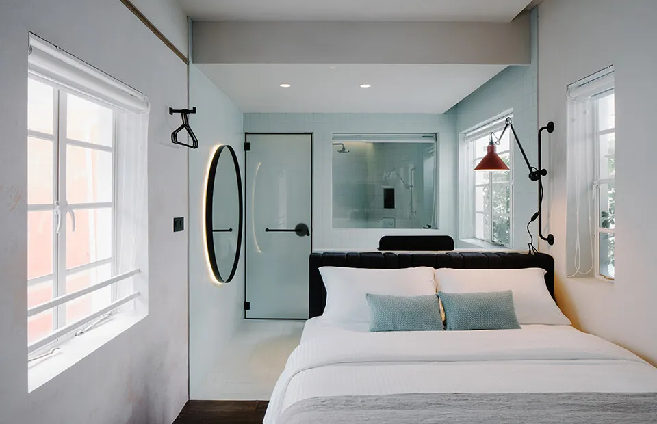 Modern minimalist bedroom featuring white concrete walls, glass shower enclosure, and a bed with white bedding and green acce