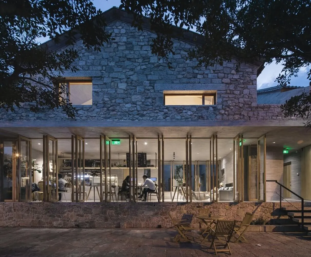 An exterior view of a stone cafe with people seated at tables inside, illuminated through large windows.