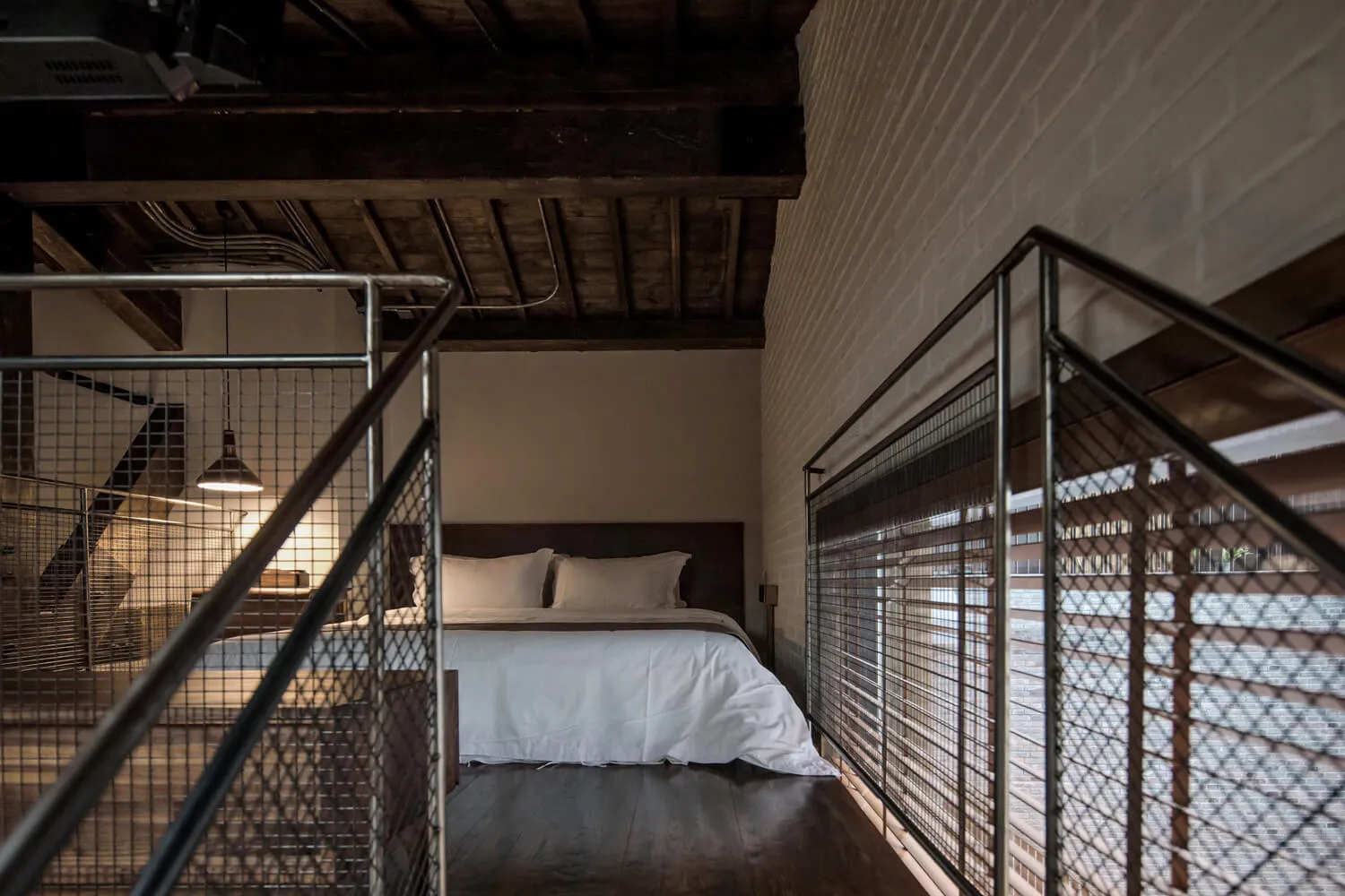 A minimalist bedroom with industrial design elements featuring exposed brick walls, wooden beam ceiling, and a metal rail.