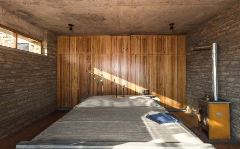 A minimalist bedroom with wooden furniture and a stone wall, featuring a concrete ceiling and a large window allowing sunligh