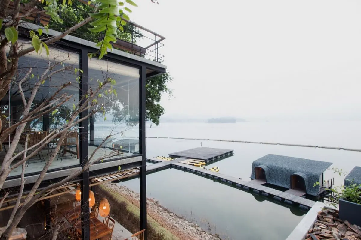Modern residential building with glass walls and wooden accents overlooking a serene water body.