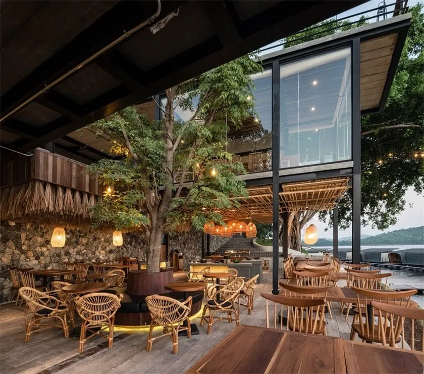 Outdoor seating area of a modern restaurant with wooden tables and chairs, glass walls, and a large tree.
