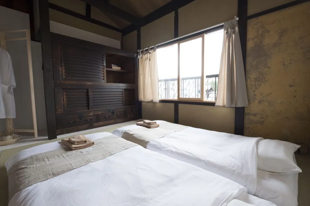 Minimalist bedroom with white bedding and wooden accents, featuring a large window with beige curtains, and a rustic wooden c