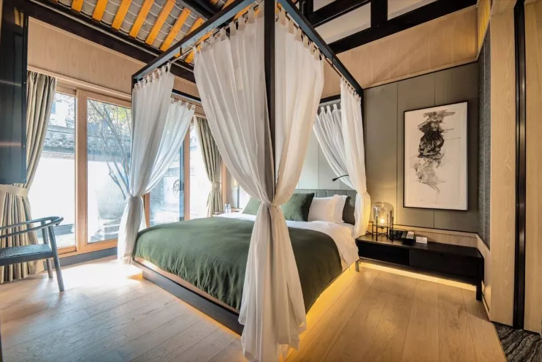 A modern minimalist bedroom featuring a canopy bed with white curtains, wooden flooring, and a black and white abstract paint