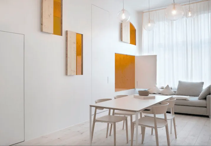 A modern, minimalist living room with a white table and chairs, light wooden flooring, white curtains, and orange accent piec