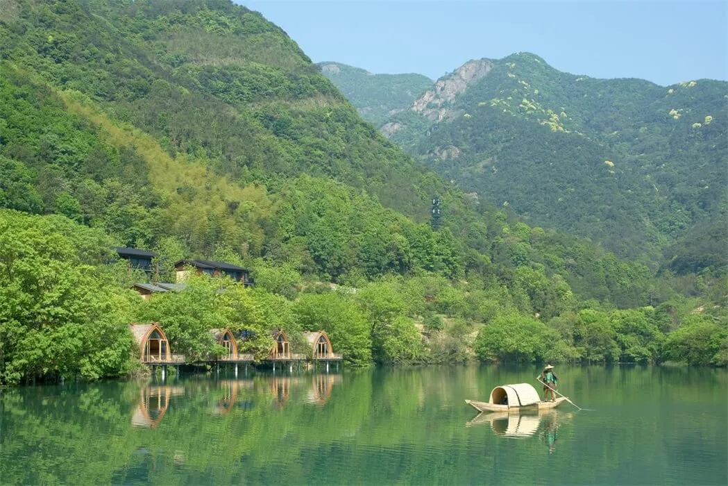 A serene waterfront resort with lush greenery, a boat, and mountains in the background.