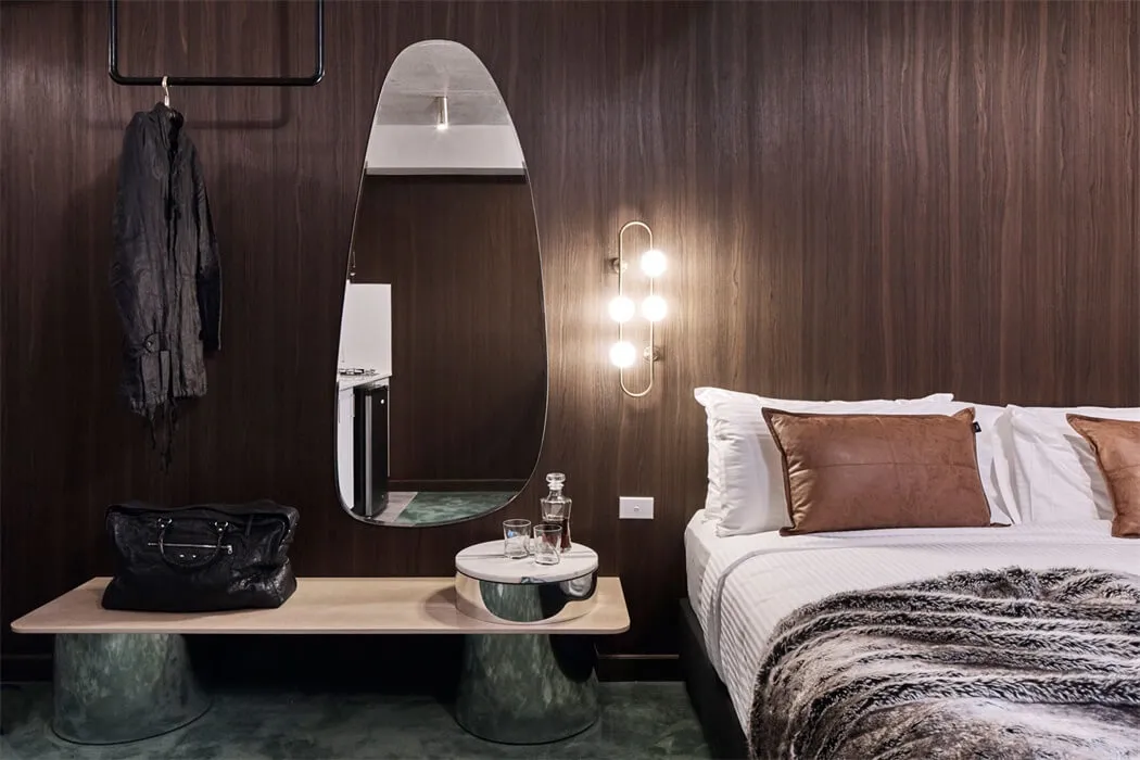 Modern bedroom featuring a wooden wall, a bed with white bedding, a round mirror, and a black leather bag on a side table.