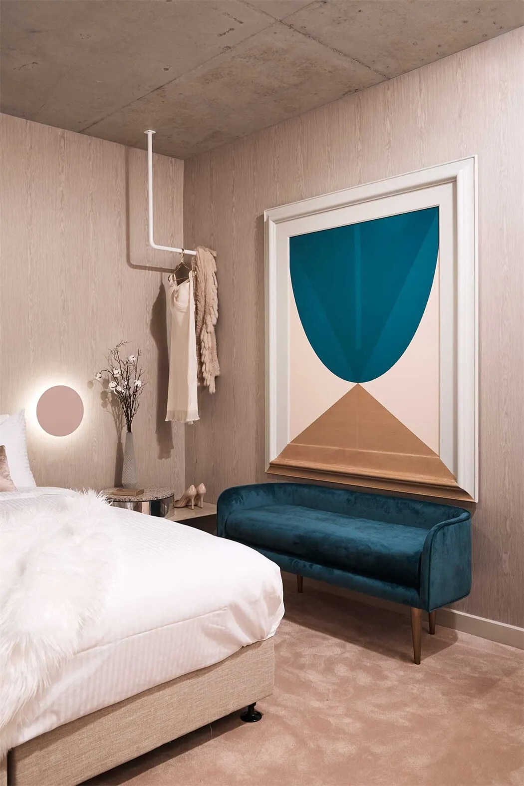 A modern minimalist bedroom with a blue velvet bench, geometric artwork, and a concrete ceiling.