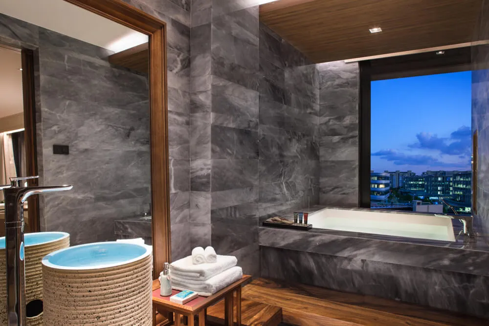 Modern minimalist bathroom featuring a marble tub, sink, and mirror with a cityscape view in the background.