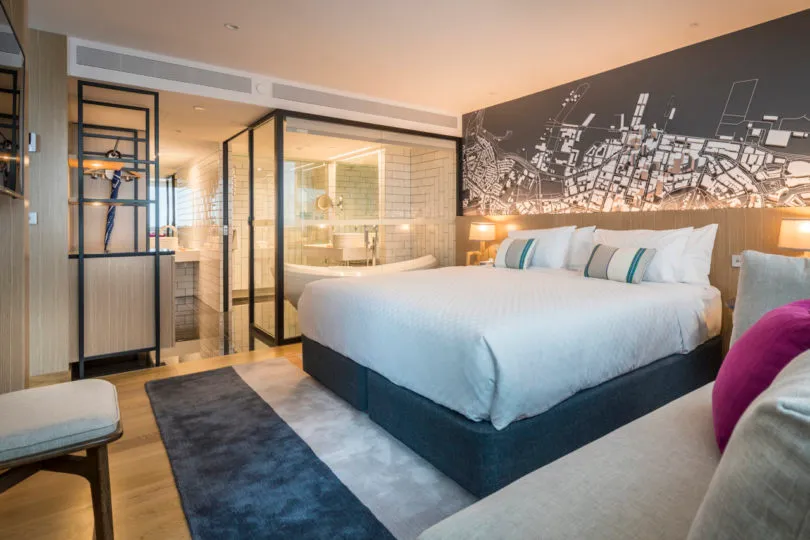 Modern bedroom featuring a neatly made bed with white linen, a comfortable seating area, and a stylish bathroom in the backgr