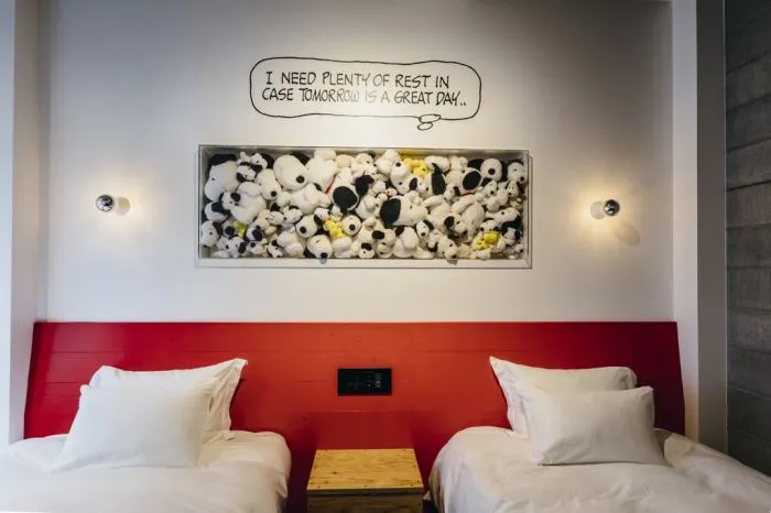 A bedroom with two beds, white pillows, a red headboard, and a wooden nightstand with a speech bubble saying, 'I need plenty