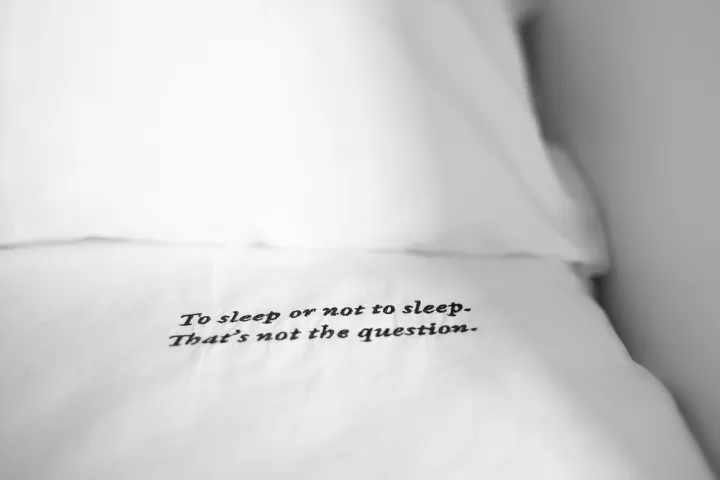 A white bed sheet embroidered with a famous Shakespearean quote, "To sleep or not to sleep. That's not the question."