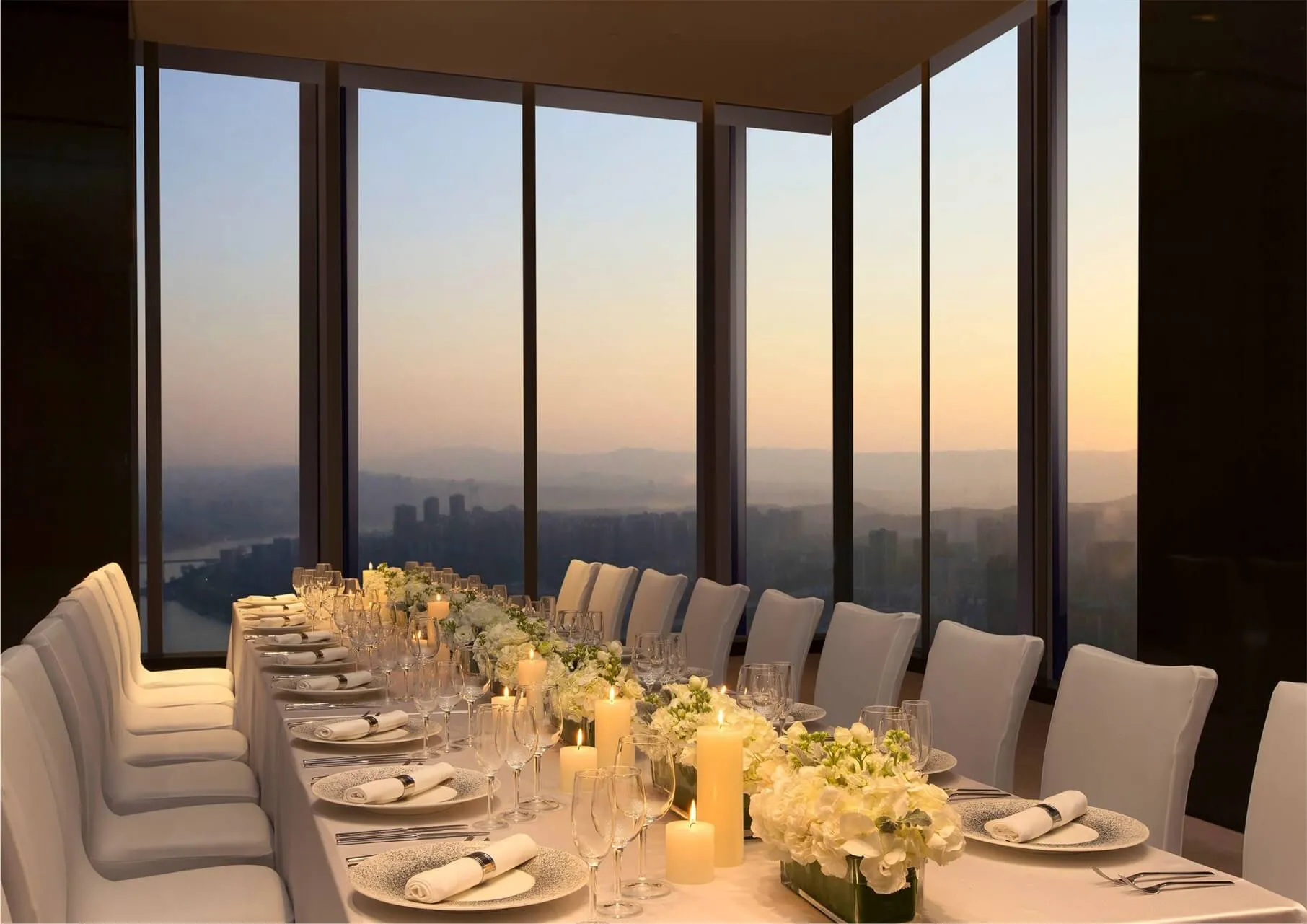 An elegant dinner party setup in a modern room with large windows showing a beautiful sunset view.
