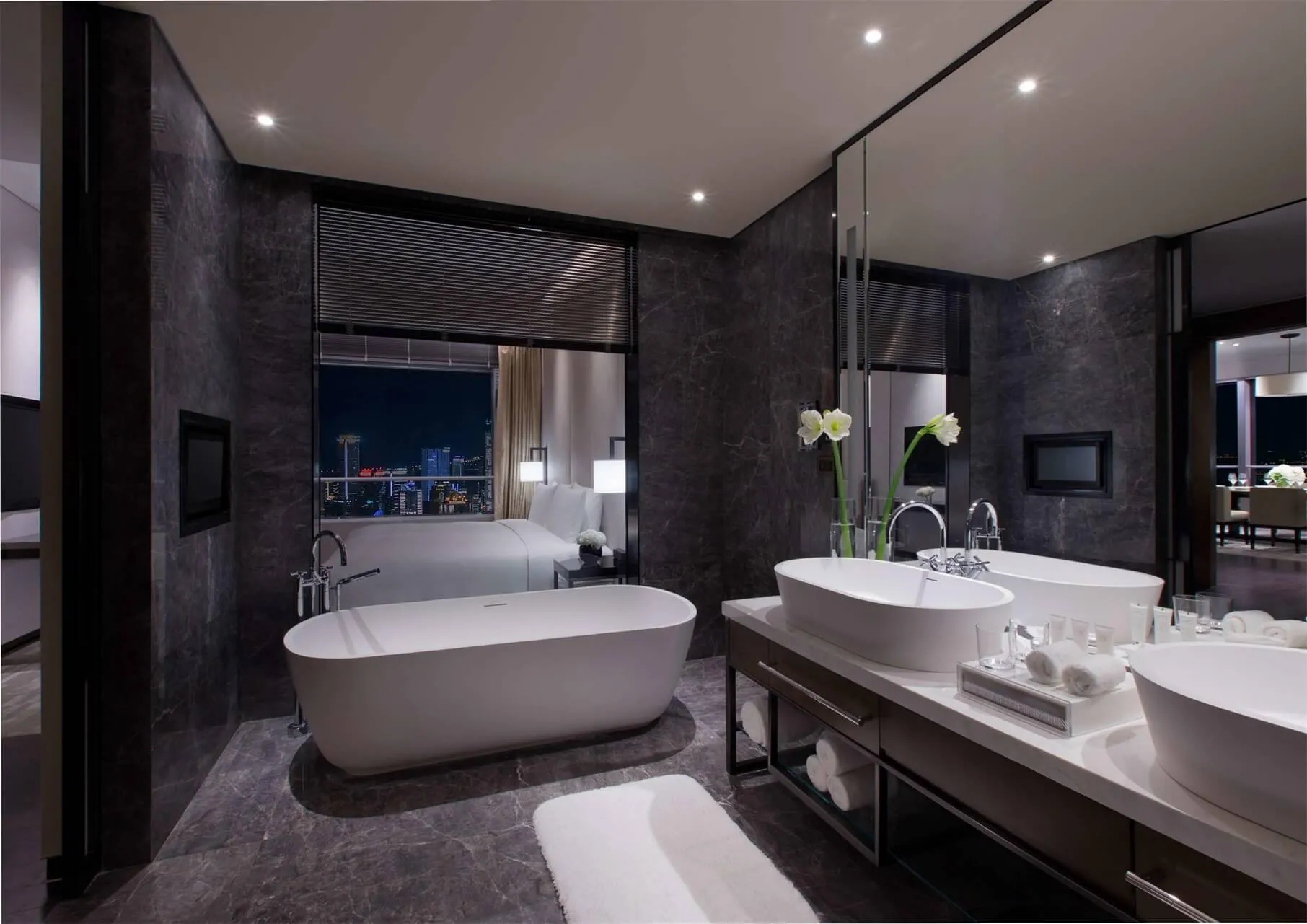 A modern bathroom with marble countertops, black granite walls, and a freestanding bathtub overlooking a cityscape.