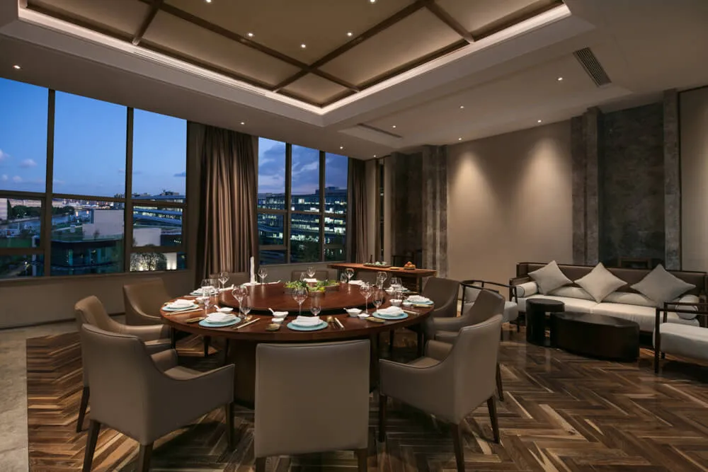 A modern residential dining room with a round wooden table, leather chairs, and large windows overlooking a cityscape.