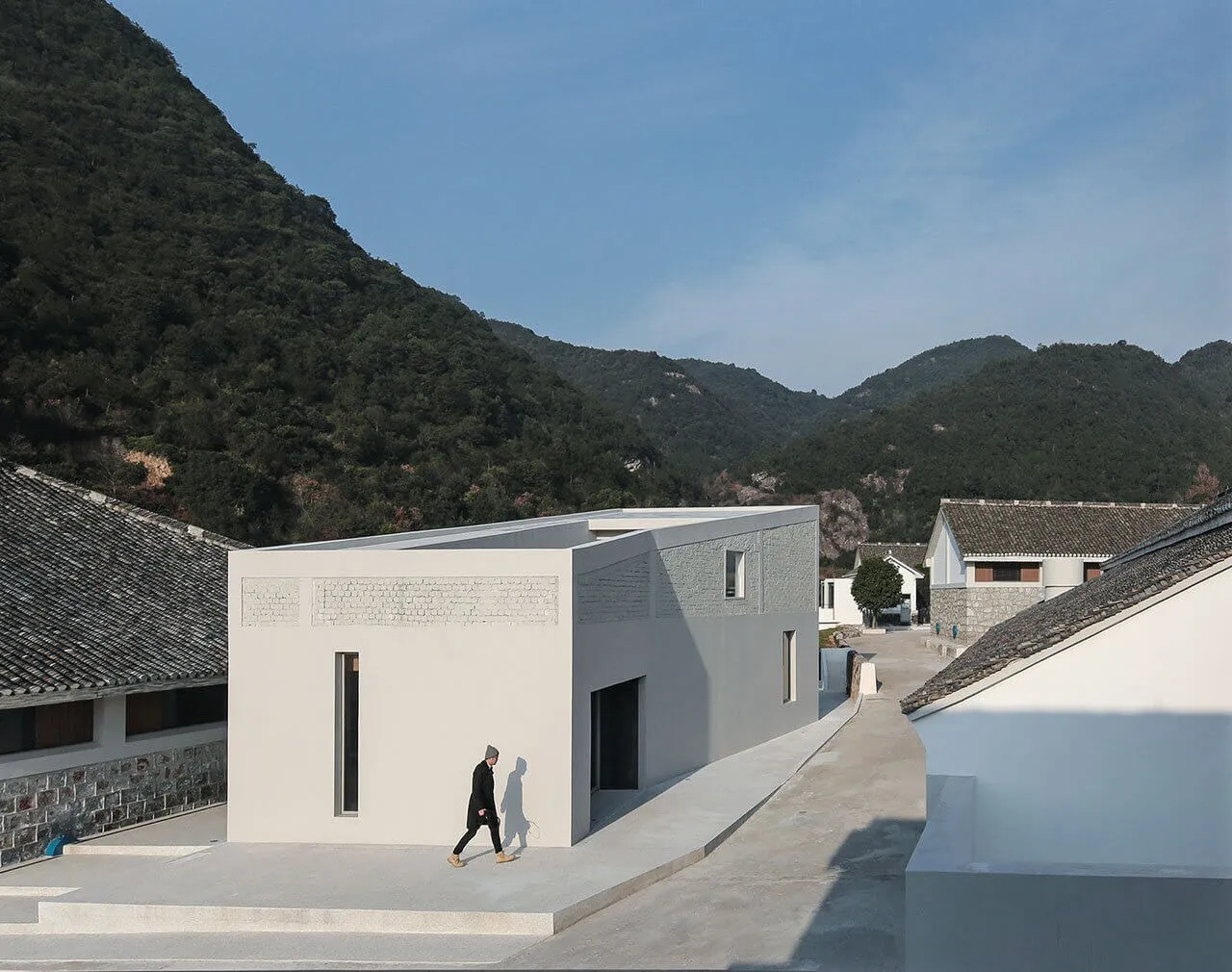 A modern building made of concrete with a person walking on a walkway in front of it, set against a backdrop of green hills a