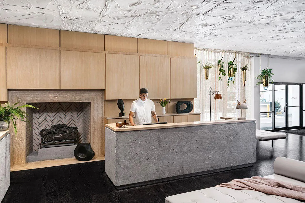 A modern, minimalist hotel reception desk made of concrete with a wooden cabinet and a cozy fireplace in the background.