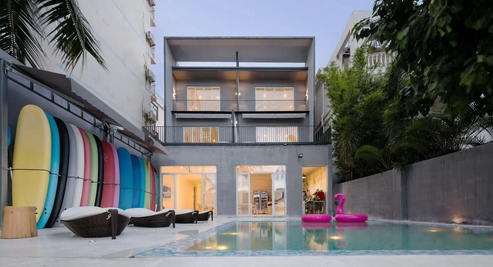 Modern minimalist house with concrete exterior, glass windows, and a pool surrounded by woven lounge chairs and colorful surf
