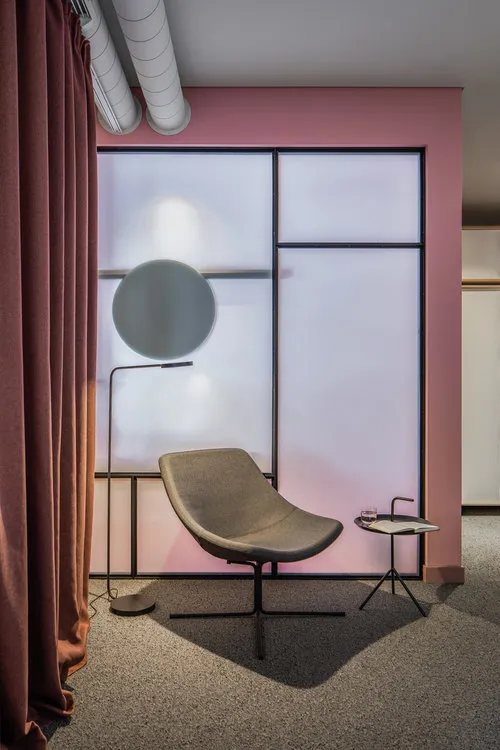 A minimalist room featuring a grey armchair, a black metal side table, a floor lamp with a white shade, and a glass partition