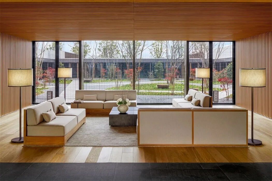 A modern living room with wood paneling, including beige sofas, a glass coffee table, and floor-to-ceiling windows overlookin