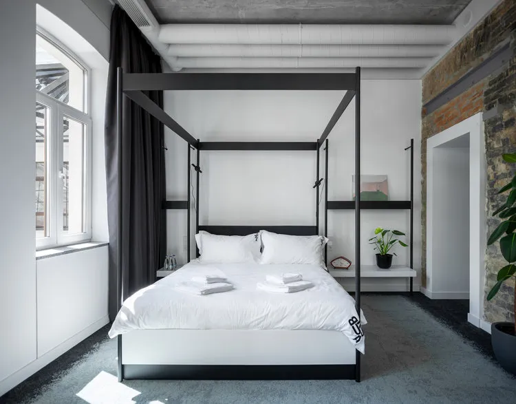 A minimalist bedroom with a concrete and metal canopy bed, white bedding, and a large window.