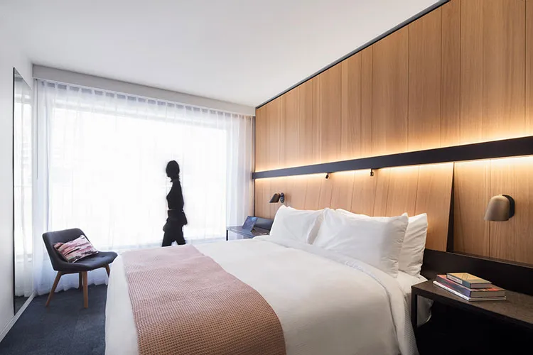 A minimalist bedroom featuring a large bed with wooden headboard, white sheets, and a side table with two books and a lamp.