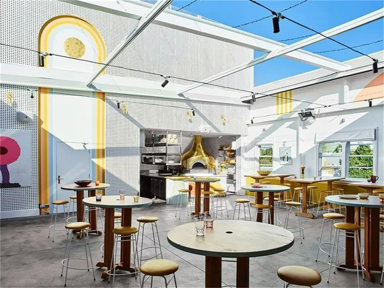 A spacious and modern cafe interior with tall bar-height tables, yellow stools, and large windows.