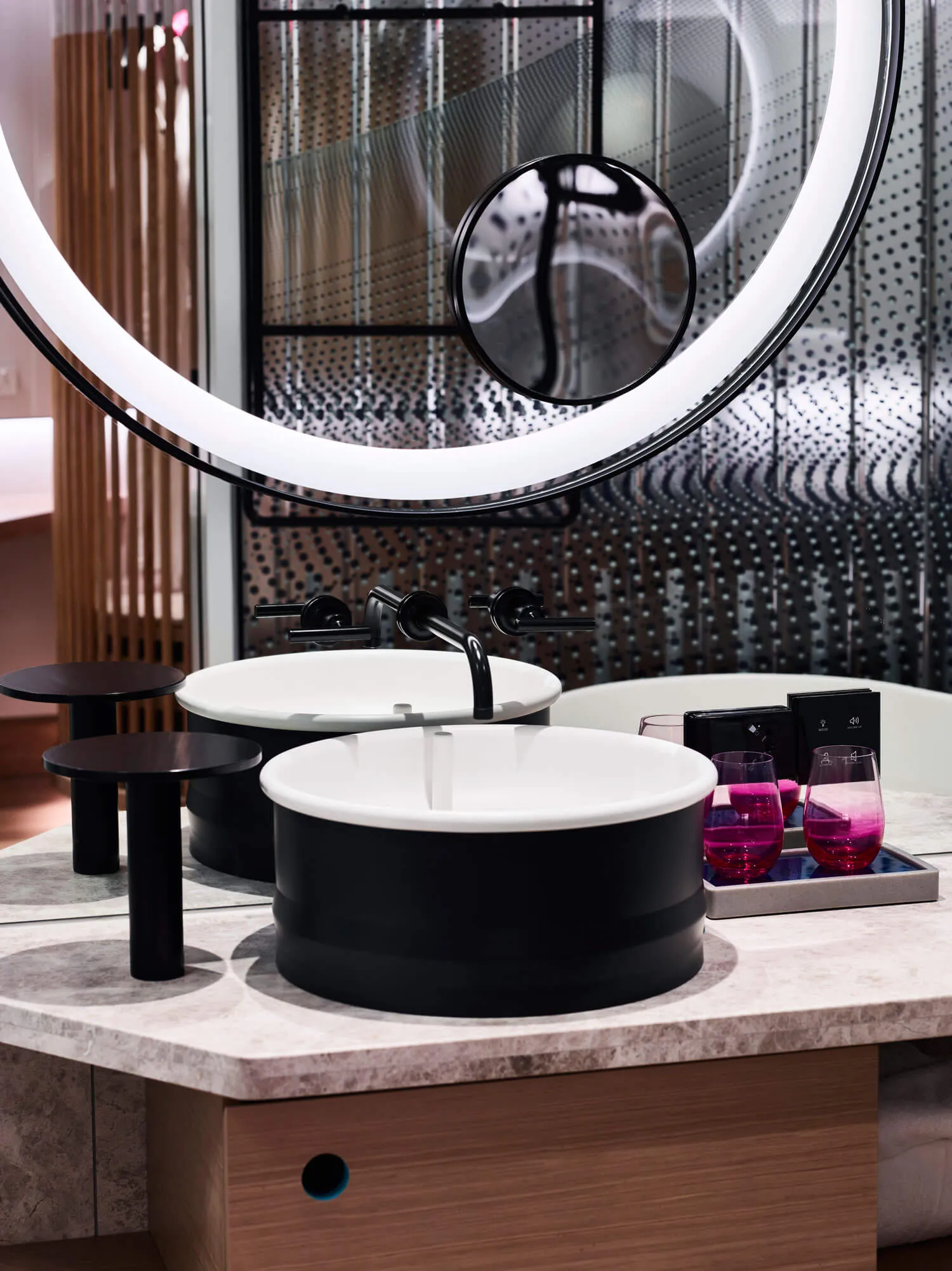 A modern bathroom featuring a sleek marble countertop with two sinks, a large round mirror, and a tray with pink glasses.