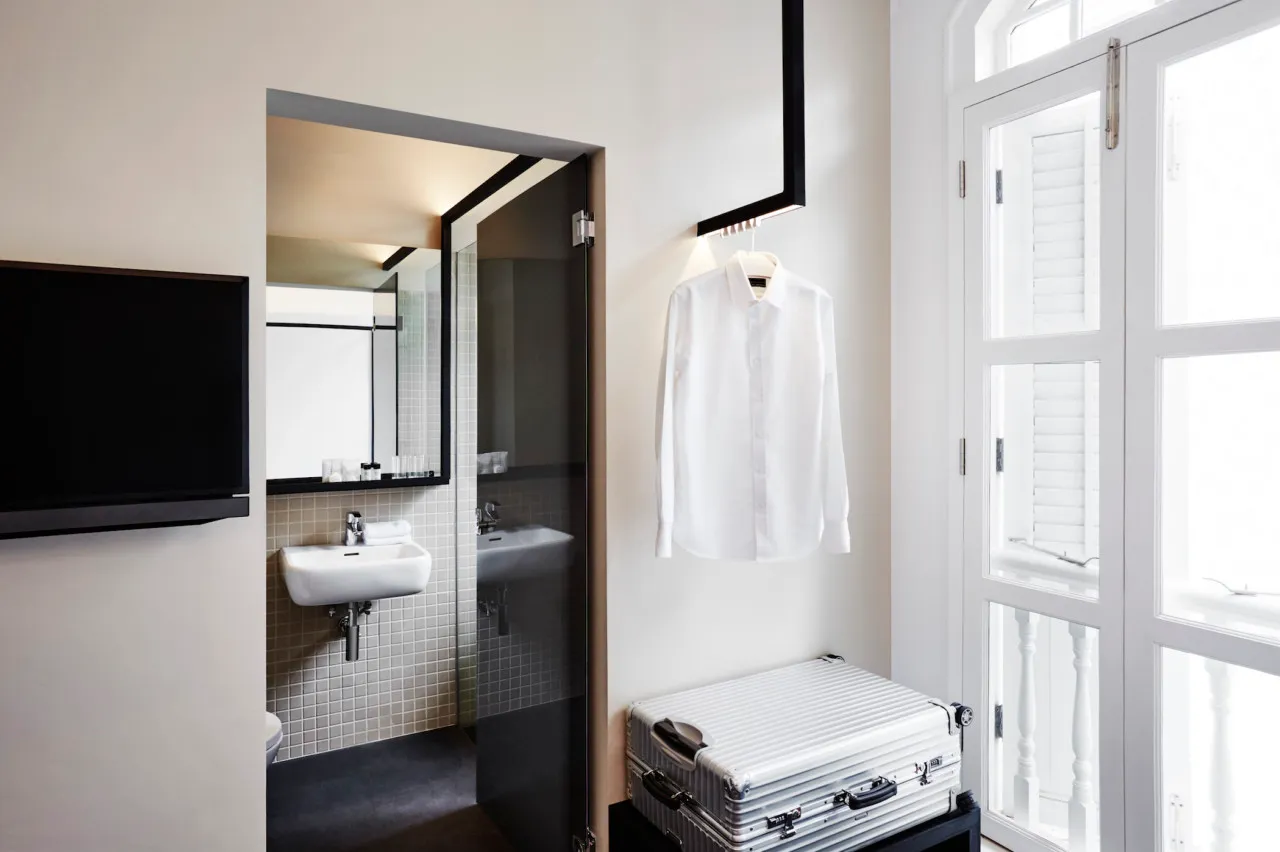 A minimalist design room in a hotel suite with a white shirt hanging on the wall, a silver suitcase, a white sink, and a tele