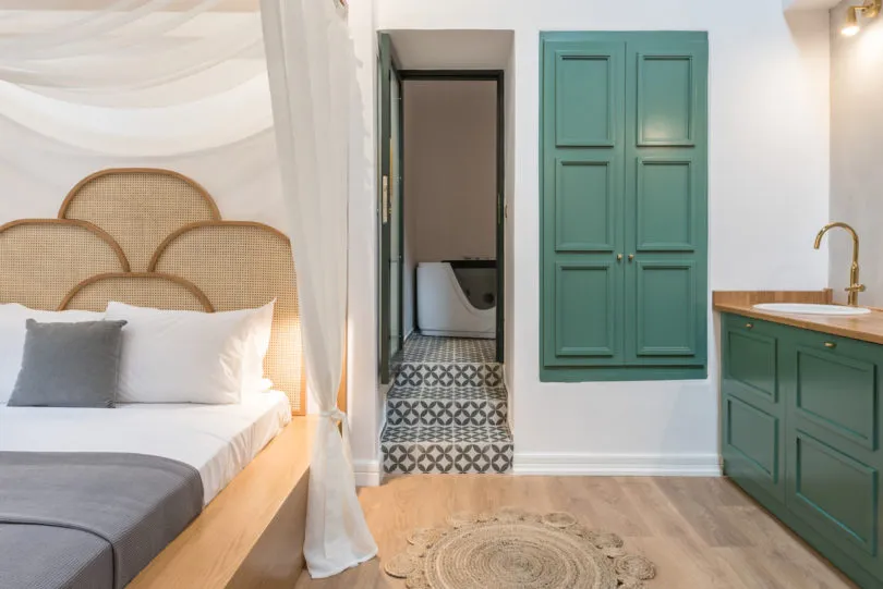 Minimalist bedroom interior featuring a cane headboard bed, wooden flooring, and green painted walls with a sink and laundry