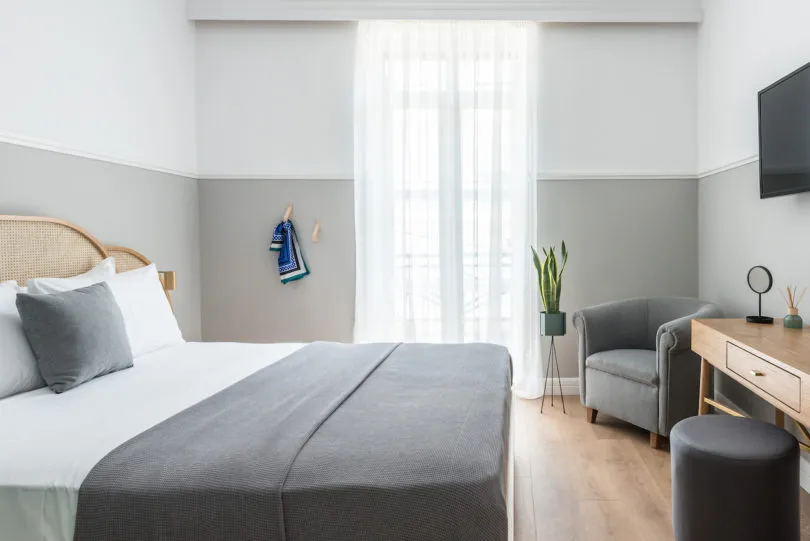 A minimalist bedroom featuring a bed with white and gray bedding, a gray armchair, a wooden desk, and a potted plant.