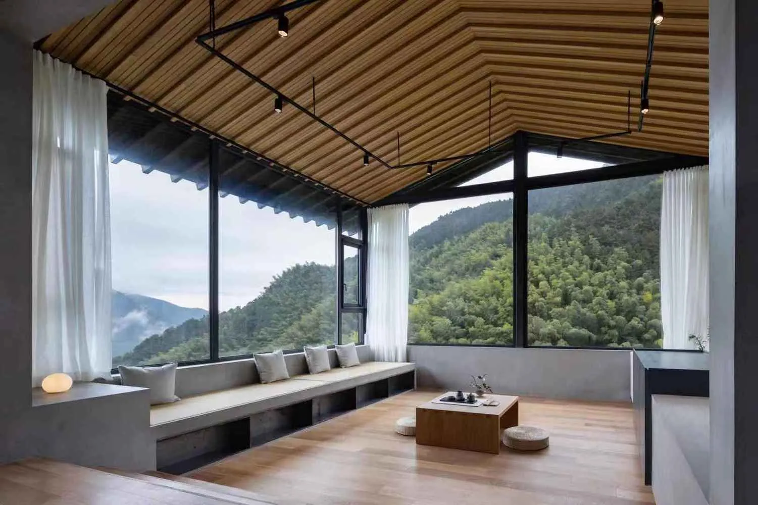 A modern living room with a wooden floor, glass windows overlooking green mountains, and concrete seating areas.