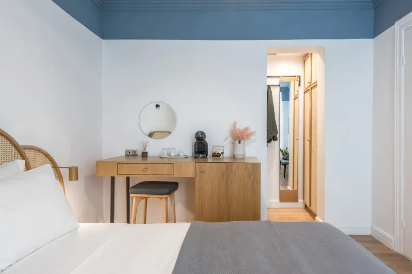 A modern minimalist bedroom featuring a wooden vanity, rattan headboard, and a large bed with white bedding and a grey blanke