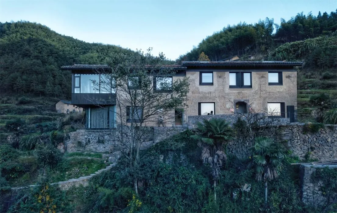 A modern residential architecture featuring a terraced stone house nestled in a lush green hillside.