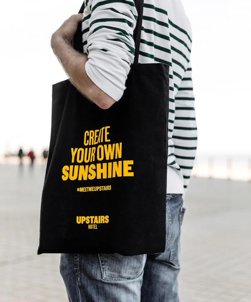 Person holding a black tote bag with 'CREATE YOUR OWN SUNSHINE' text on it.