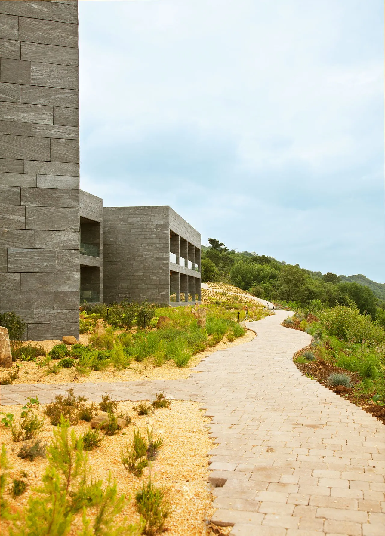Modern landscape architecture featuring stone structures, gravel pathways, and various plants in a natural setting.