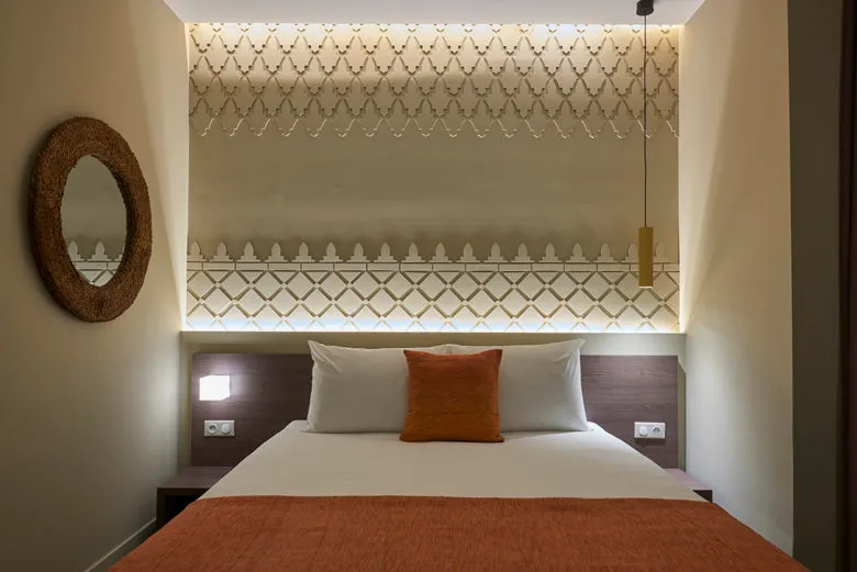 Modern bathroom with white and orange bedding on a bed, textured white walls, and a round mirror.