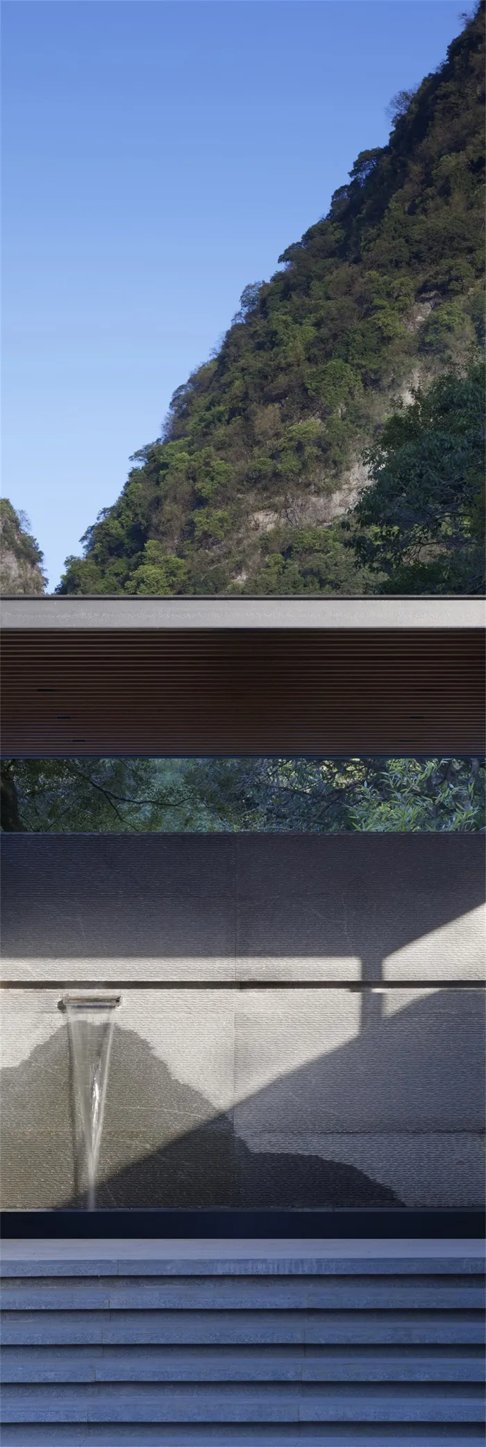 A modern architectural design featuring concrete steps and a concrete wall with a water feature, set against a natural mounta