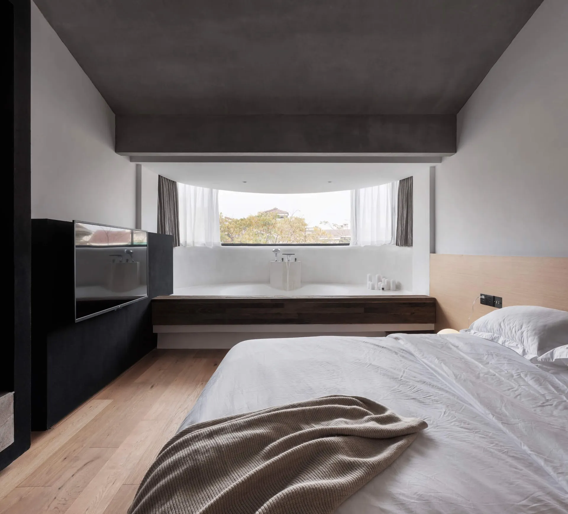 A modern minimalist bedroom with concrete walls, wood flooring, and a large window.